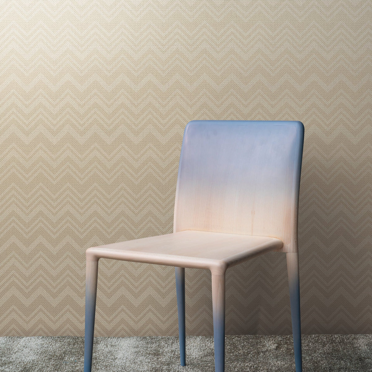 A modern chair with a gradient color from blue at the top to beige at the bottom is set against a wall covered in York Wallcoverings’ Iconic Shades Wallpaper featuring a zigzag pattern, complemented by a textured gray carpet.