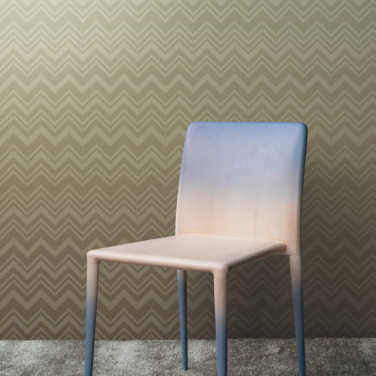 A chair with a gradient blue and beige color sits on a gray carpet in front of a tan Zigzag-patterned Iconic Shades Wallpaper by York Wallcoverings.