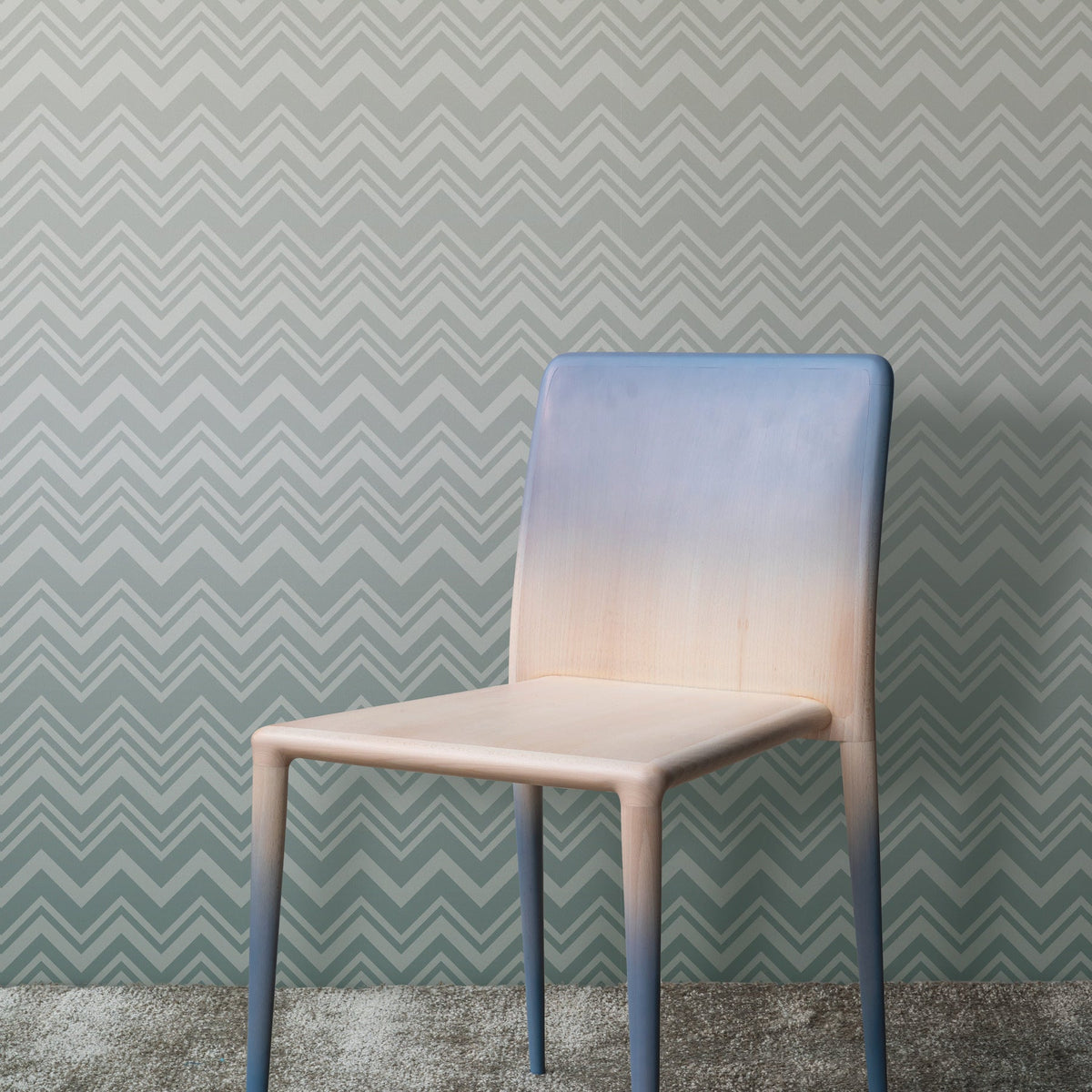 A wooden chair with a gradient blue and beige finish stands on a gray carpet in front of the Iconic Shades Wallpaper by York Wallcoverings, which features a gray zigzag pattern.