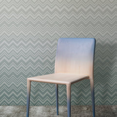 A wooden chair with a gradient blue and beige finish stands on a gray carpet in front of the Iconic Shades Wallpaper by York Wallcoverings, which features a gray zigzag pattern.