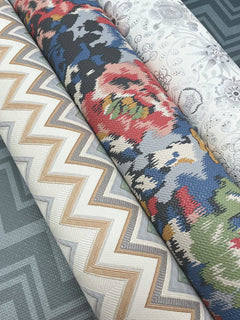 Three rolls of York Wallcoverings’ Iconic Shades Wallpaper—one featuring a gold zigzag pattern, another showcasing a colorful floral design, and the third displaying a white lace-like floral motif—are arranged on a blue geometric-patterned background.