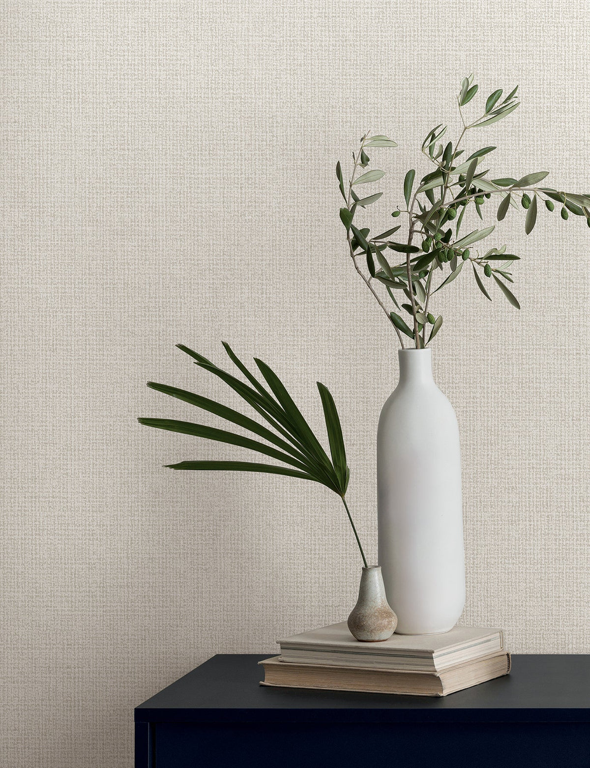 Two white vases containing greenery rest on a stack of books against textured Magnolia Home Brindled Wallpaper.