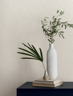 Two white vases containing greenery rest on a stack of books against textured Magnolia Home Brindled Wallpaper.