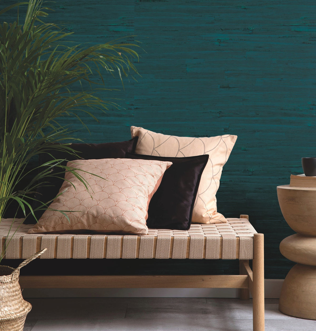 A woven wooden bench with beige and black cushions sits beside a round wooden side table. A leafy plant and a woven basket are nearby, with York Wallcoverings’ Ronald Redding Lotus Leaf Wallpaper in teal textured tones featured on the wall in the background.