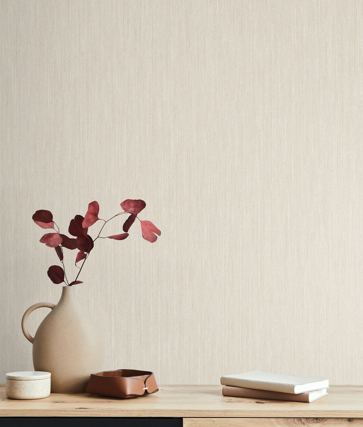 Smooth as Silk Wallpaper by York Wallcoverings in light beige with a subtle vertical texture, featuring a vase with reddish-brown leaves and small personal items on a wooden surface in the foreground.