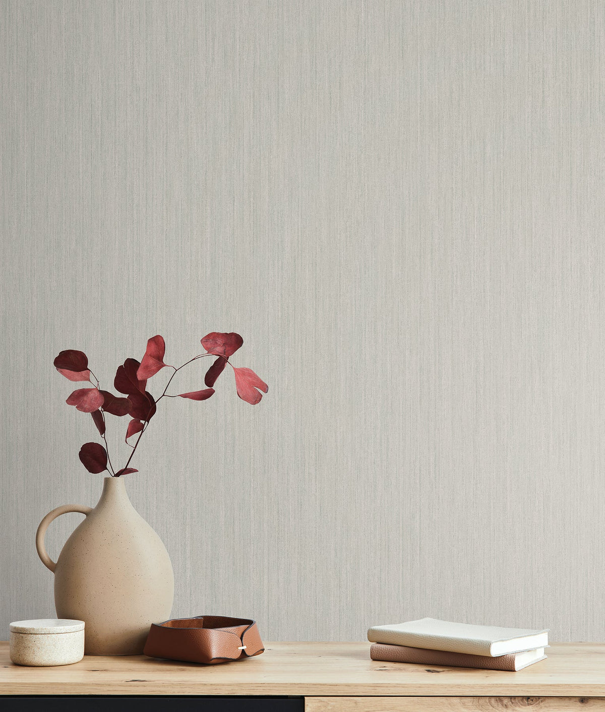 Smooth as Silk Wallpaper by York Wallcoverings serves as a light gray textured backdrop for a wooden surface holding a vase of red leaves, a small container, a bowl, and two stacked books.