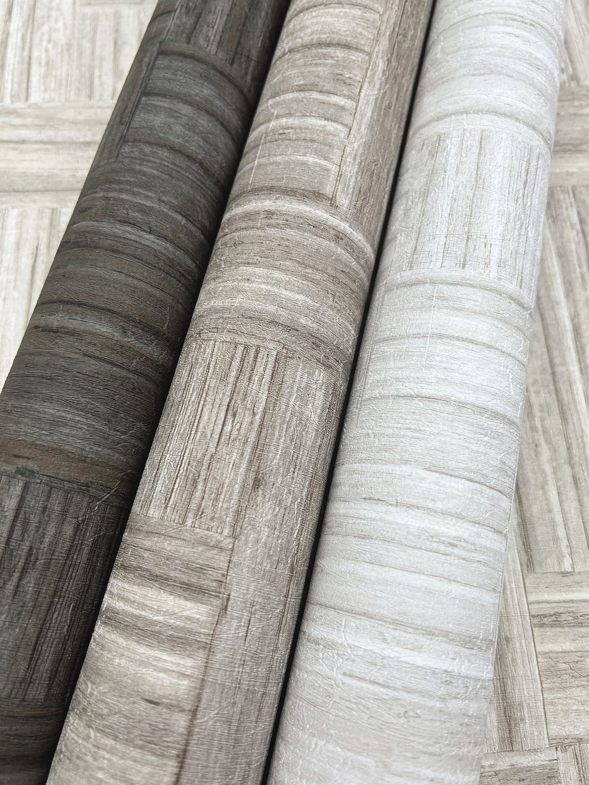 Three rolls of Tesselle Wallpaper featuring a geometric wood-grain pattern in charcoal, taupe, and off-white.