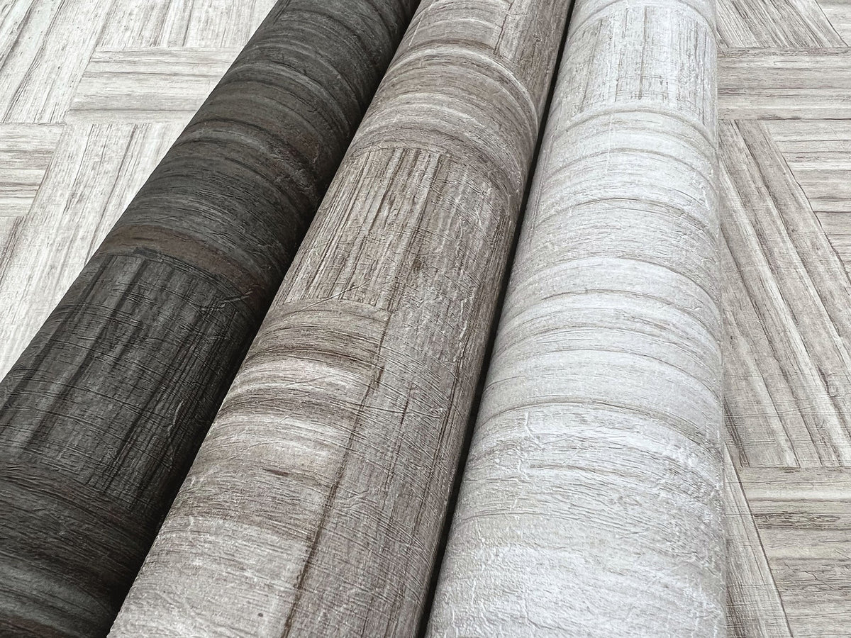 Three rolls of Tesselle Wallpaper featuring a wood-grain pattern in charcoal, brown, and light gray, displayed against a background of the same pattern.