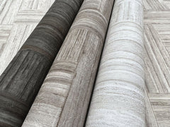 Three rolls of Tesselle Wallpaper featuring a wood-grain pattern in charcoal, brown, and light gray, displayed against a background of the same pattern.