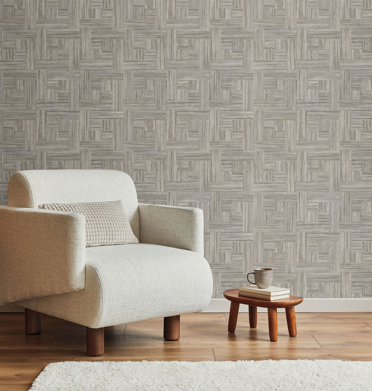 A neutral armchair sits next to a small wooden stool with books and a mug, set against York Wallcoverings Tesselle Wallpaper featuring a repeating geometric wood-grain pattern.