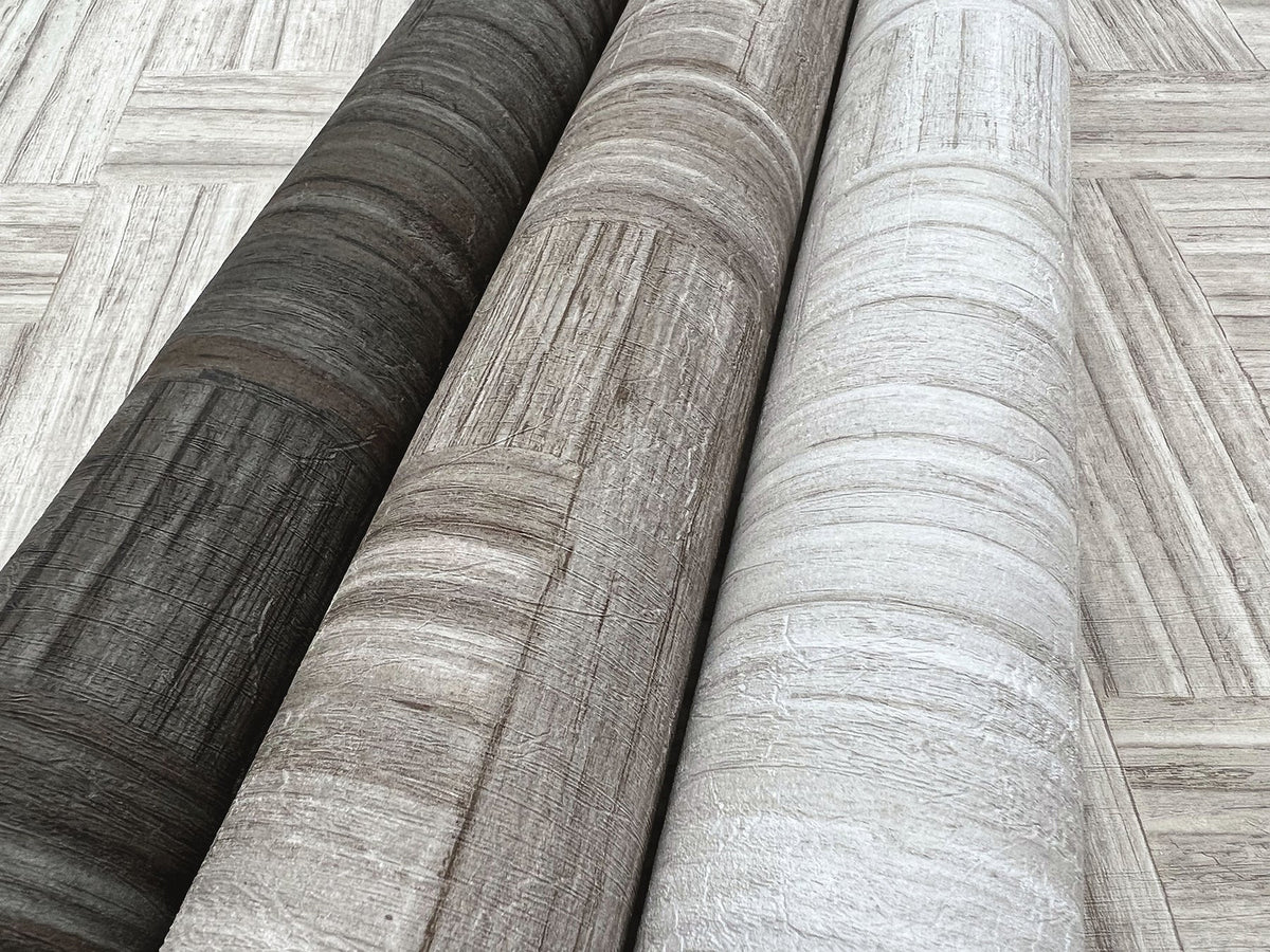 Three rolls of Tesselle Wallpaper in dark charcoal, light brown, and off-white featuring a textured wood-grain pattern, lying on a surface with the same geometric wood-plank design.