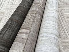 Three rolls of Tesselle Wallpaper in dark charcoal, light brown, and off-white featuring a textured wood-grain pattern, lying on a surface with the same geometric wood-plank design.