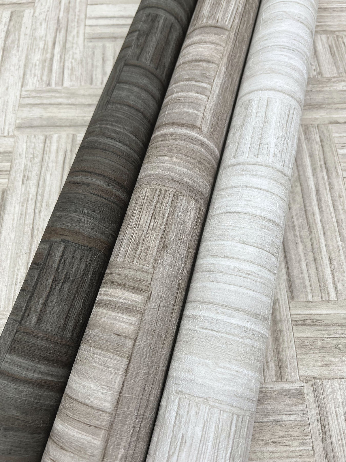 Three rolls of Tesselle Wallpaper by York Wallcoverings featuring a wood-grain geometric pattern in charcoal, taupe, and white, resting on a matching surface.