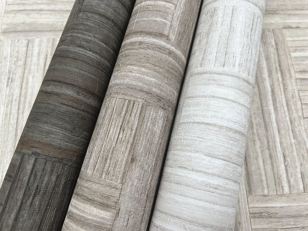 Three rolls of Tesselle Wallpaper featuring a wood-grain geometric pattern in shades of charcoal, taupe, and light gray.