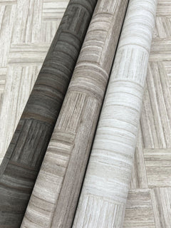 Three rolls of Tesselle Wallpaper featuring a wood-look geometric tile pattern in charcoal, taupe, and cream, resting on a matching surface.