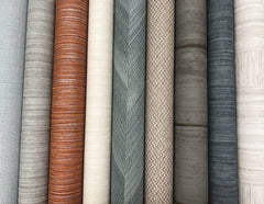 Ten rolls of Tasar Silk Wallpaper by York Wallcoverings, in a range of neutral shades such as gray, brown, beige, olive, and cream, are arranged vertically side by side. The collection features various patterns including solid weaves, chevrons, and stripes.