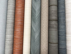 Rolls of York Wallcoverings’ Tasar Silk Wallpaper in a variety of colors and textures—including shades of grey, beige, brown, green, and orange—are lined up vertically side by side. Each roll features its own unique pattern or weave.