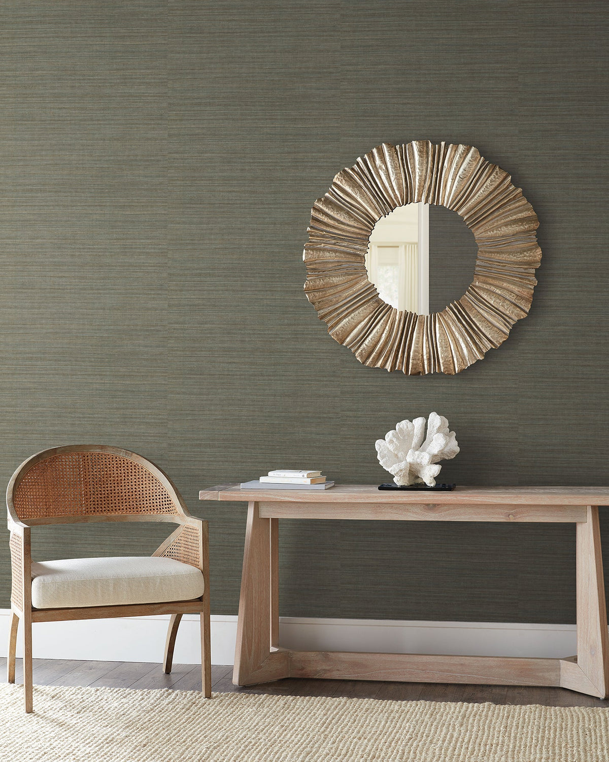 Dark, textured Tasar Silk Wallpaper by York Wallcoverings behind a wooden console table with a coral sculpture, a circular sunburst mirror, and a cane-backed chair.
