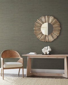 Dark, textured Tasar Silk Wallpaper by York Wallcoverings behind a wooden console table with a coral sculpture, a circular sunburst mirror, and a cane-backed chair.