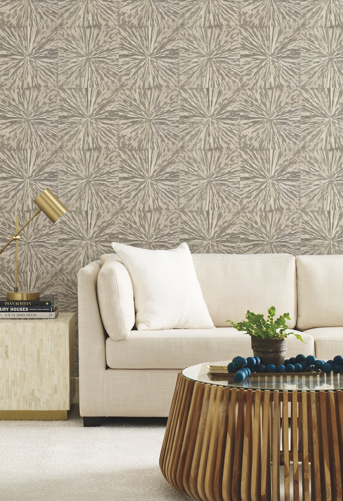 Modern living room with a beige sofa, white pillow, gold lamp on a side table, round wooden coffee table with decor, and York Wallcoverings’ Squareburst Wallpaper in neutral tones.