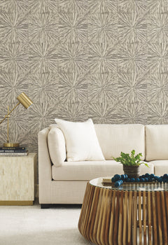 Modern living room with a beige sofa, white pillow, gold lamp on a side table, and a wooden coffee table adorned with beads and a green plant. The wall features Squareburst Wallpaper in neutral tones by York Wallcoverings.