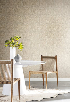 Two wooden chairs with woven seats surround a white round table. A white vase with green leaves and a small gray vase rest on the tabletop. The background features York Wallcoverings’ Squareburst Wallpaper in a beige geometric pattern, paired with a fur rug underneath.