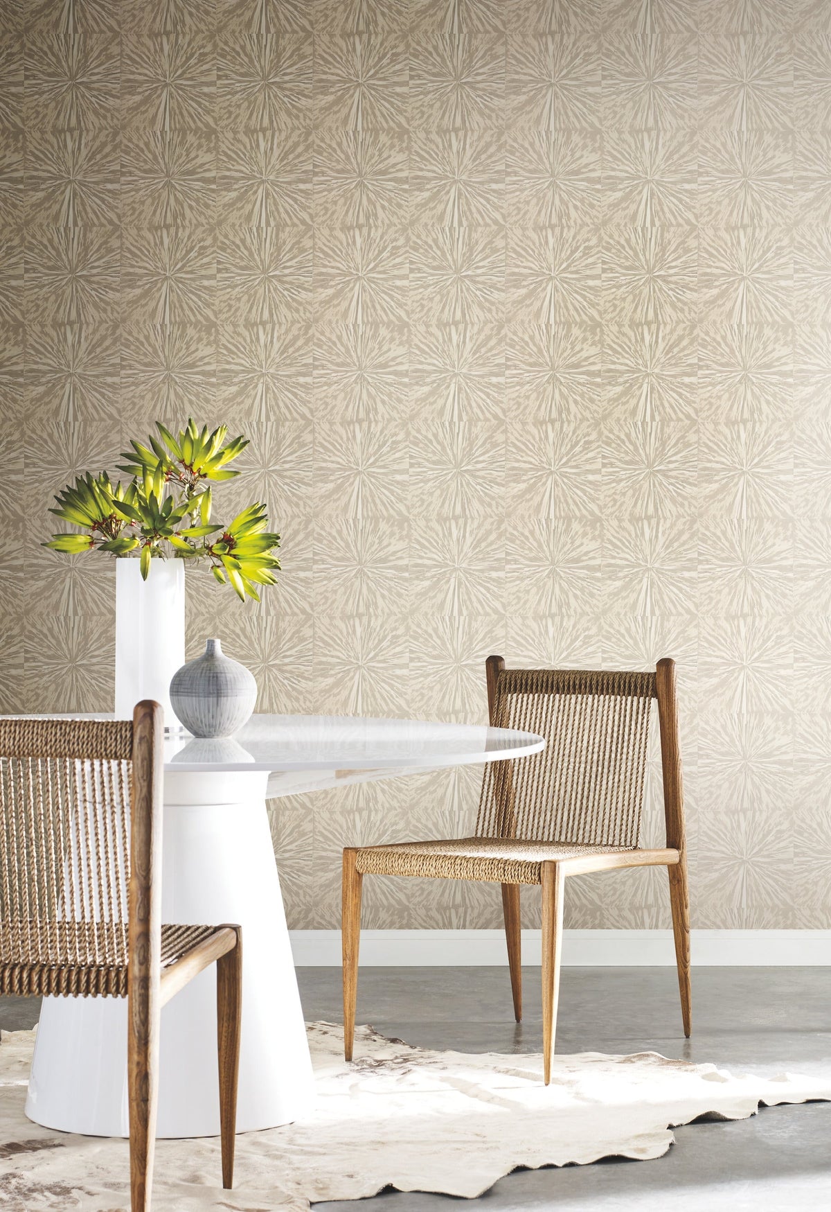 A modern dining space featuring a round white table, two woven wooden chairs, a gray vase, and a white vase with green leaves, arranged on a cowhide rug against a York Wallcoverings Squareburst Wallpaper accent wall.