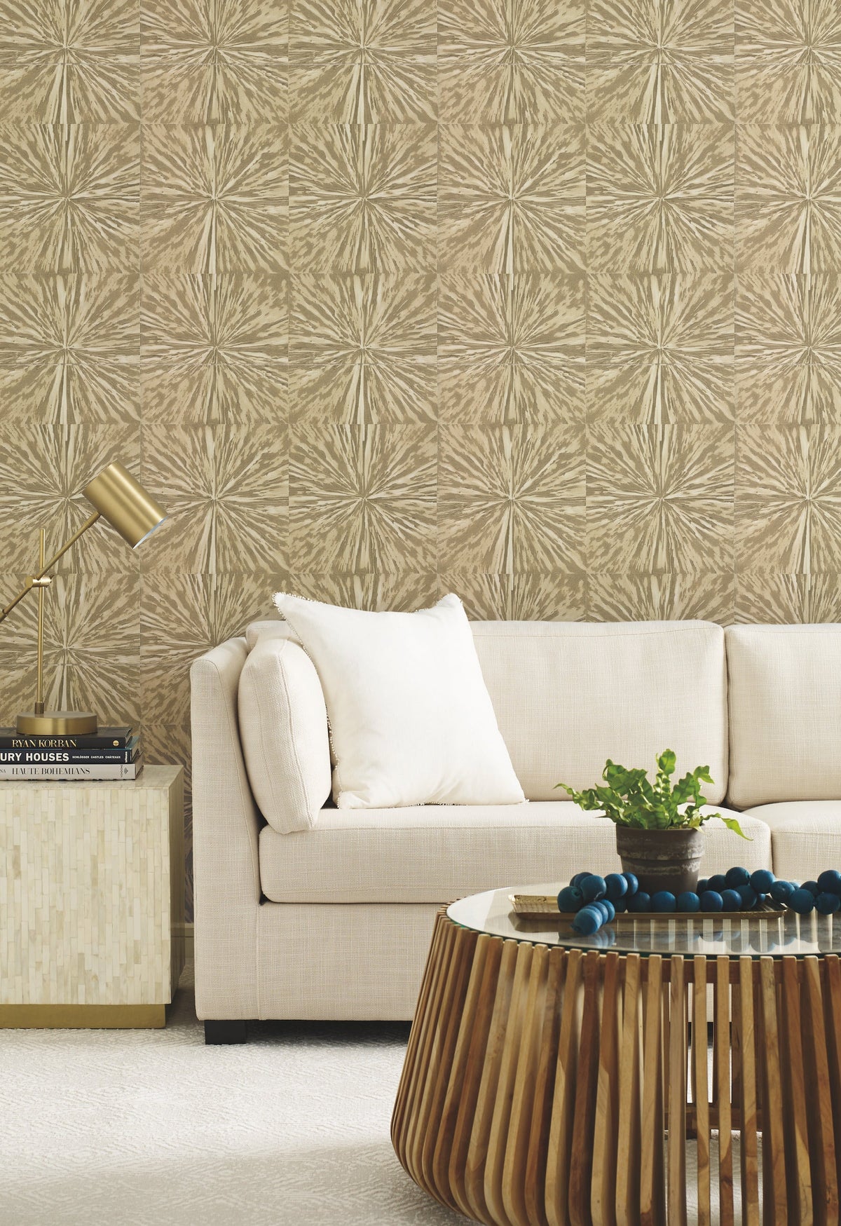A modern living room featuring a light beige sofa, a cushion, a small table with a gold lamp, a round wooden coffee table, a plant, and York Wallcoverings’ Squareburst Wallpaper in a geometric beige pattern.