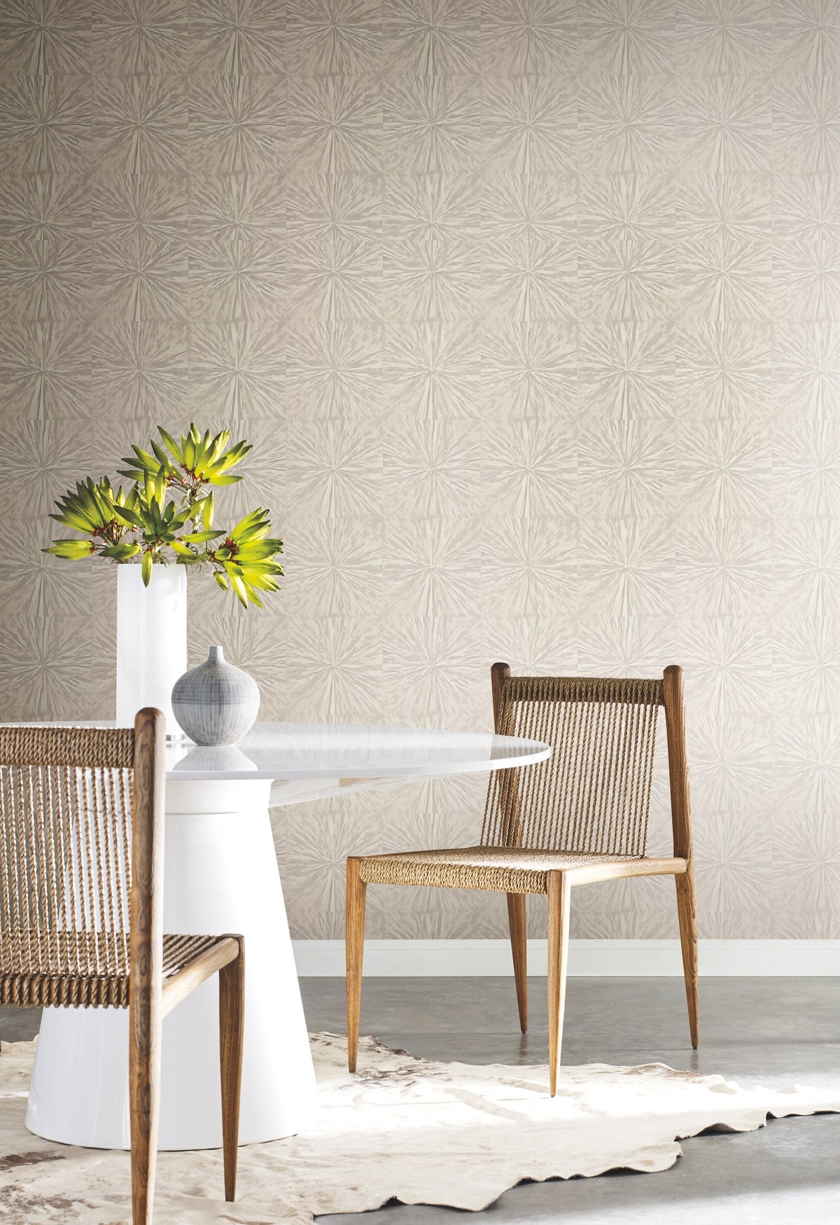 A modern dining area with a round white table, two wooden chairs featuring woven seats, a gray vase, a white vase with green foliage, and York Wallcoverings’ Squareburst Wallpaper adding a light pattern to the walls. A cowhide rug lies beneath the table.