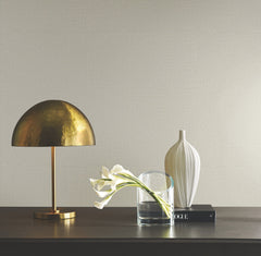 A brass dome lamp, a clear glass with calla lilies, a ribbed white vase, and a book sit on a dark wooden surface against a wall covered in Ronald Redding Tatami Weave Wallpaper by York Wallcoverings.