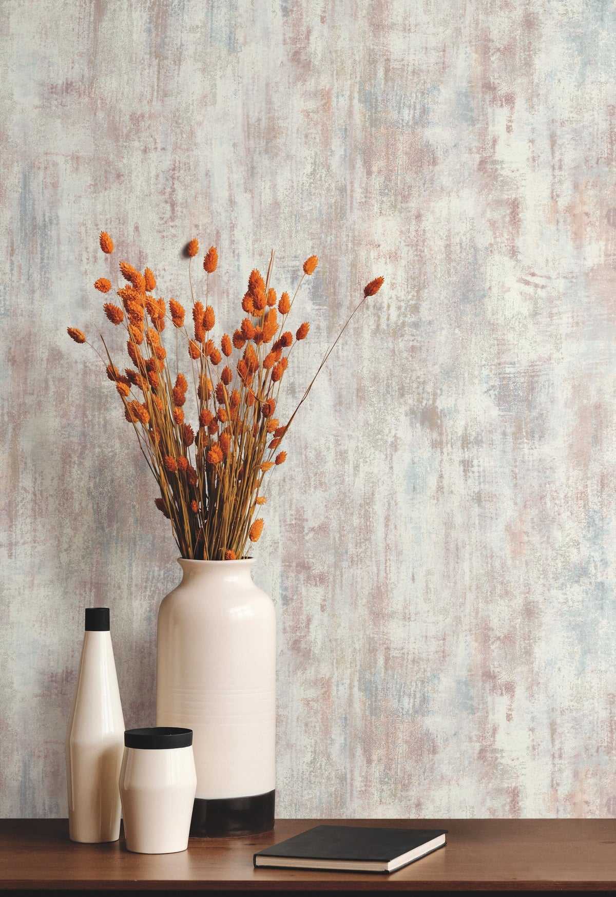 Minimalist decor featuring a large white vase with orange dried flowers, two smaller vases, and a closed black notebook placed on a wooden surface, set against York Wallcoverings’ Concrete Patina Wallpaper.