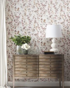Candice Olson Joyful Eucalyptus Wallpaper featuring a watercolor pattern of delicate eucalyptus branches in muted pink and green tones, shown behind a wooden dresser topped with a lamp and vases of greenery and white flowers.