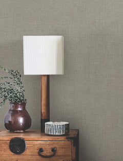 A white pleated lampshade on a wooden lamp, a brown vase with greenery, and a small striped bowl sit on a wooden chest against a wall covered with Hardy Linen Wallpaper by York Wallcoverings.