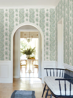 A hallway adorned with Beaumont Floral Stripe Wallpaper by York Wallcoverings in green and white, featuring a black bench with a cushion, and an arched doorway opening into a bright dining area with a round table, chairs, fresh flowers, and modern light fixtures.