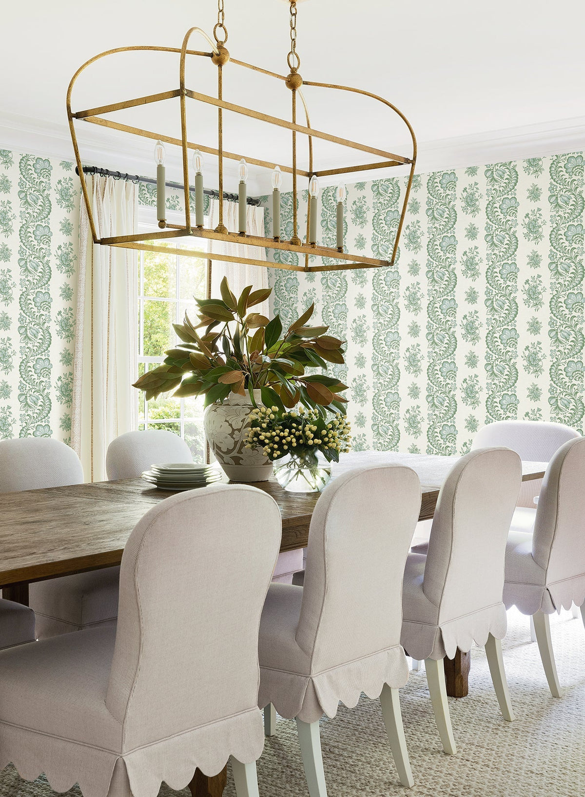 A bright dining room featuring a wooden table, white scalloped chairs, a large gold chandelier, York Wallcoverings’ Beaumont Floral Stripe Wallpaper, and a vase filled with green and white flowers as the centerpiece.