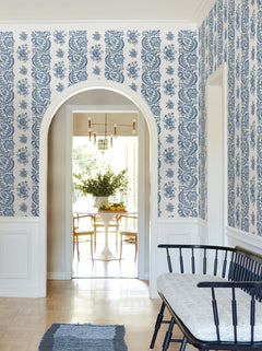 An arched doorway opens to a bright dining area with a white table and chairs. The hallway features Beaumont Floral Stripe Wallpaper by York Wallcoverings in blue and white, along with a black bench topped with a cushion and a blue rug on light wood floors.