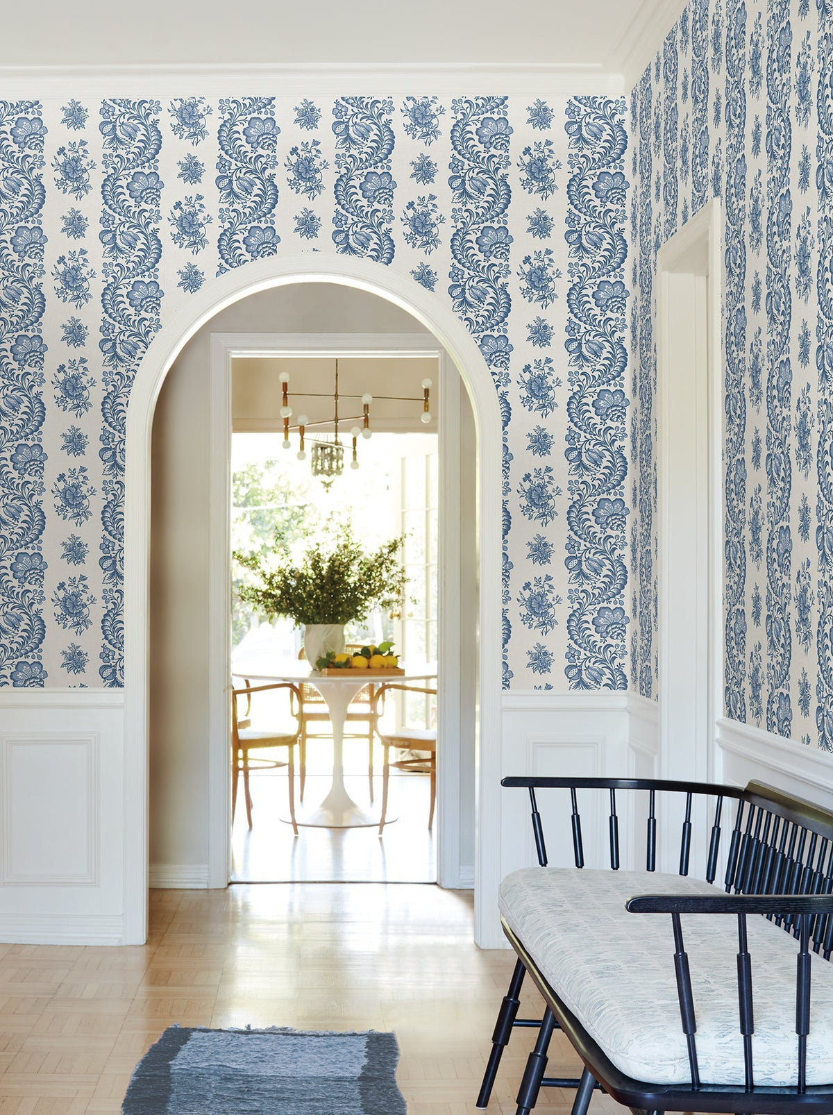 An arched doorway leads to a bright dining area with a round table and chairs. The walls are adorned with Beaumont Floral Stripe Wallpaper by York Wallcoverings, while a black bench with a cushion sits in the hallway.
