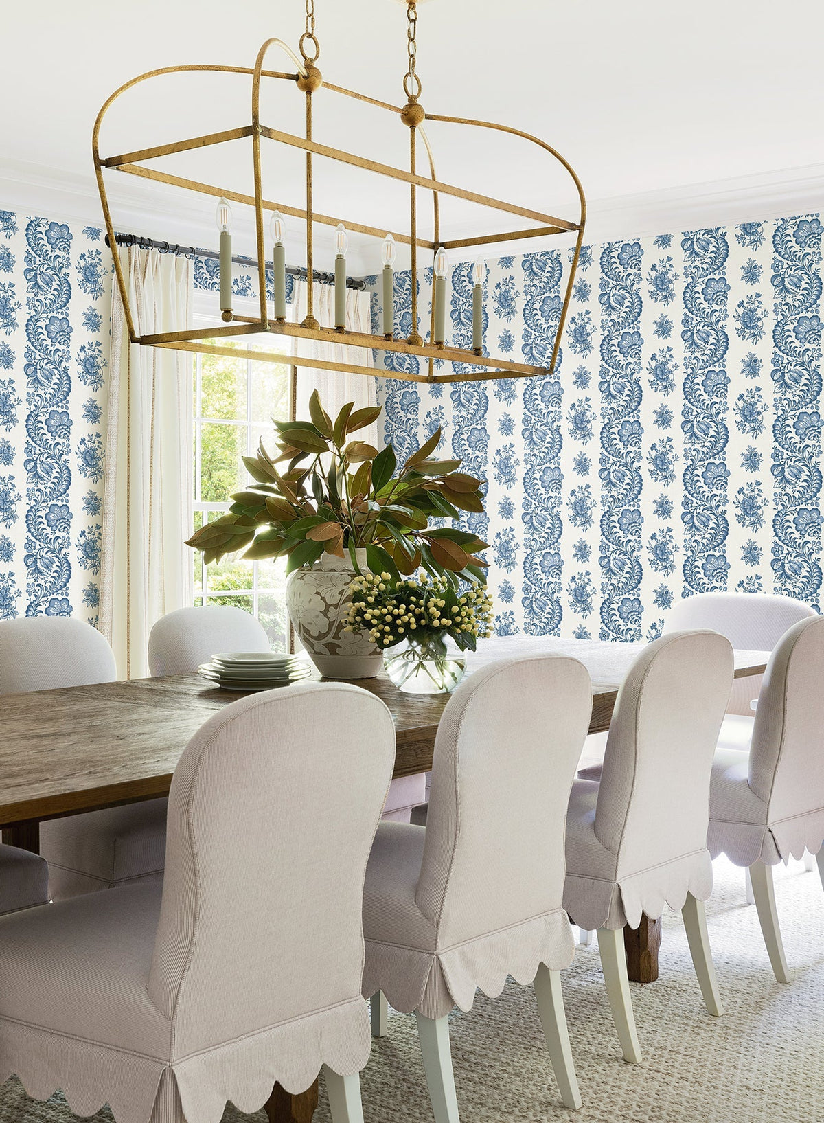A bright dining room featuring a wooden table, white scalloped chairs, a gold chandelier, and a floral centerpiece is accented by York Wallcoverings’ Beaumont Floral Stripe Wallpaper in blue and white. Sunlight streams through the window adorned with white curtains.