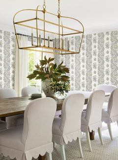 A bright dining room featuring a wooden table, eight white upholstered chairs, a gold rectangular chandelier, York Wallcoverings’ Beaumont Floral Stripe Wallpaper on the walls, and a vase of leafy branches and flowers as the centerpiece.