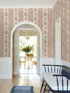 A hallway adorned with York Wallcoverings’ Beaumont Floral Stripe Wallpaper features an arched doorway, a black bench with a cushion, and offers a view into a dining room with a round table, chairs, and a vase of flowers.