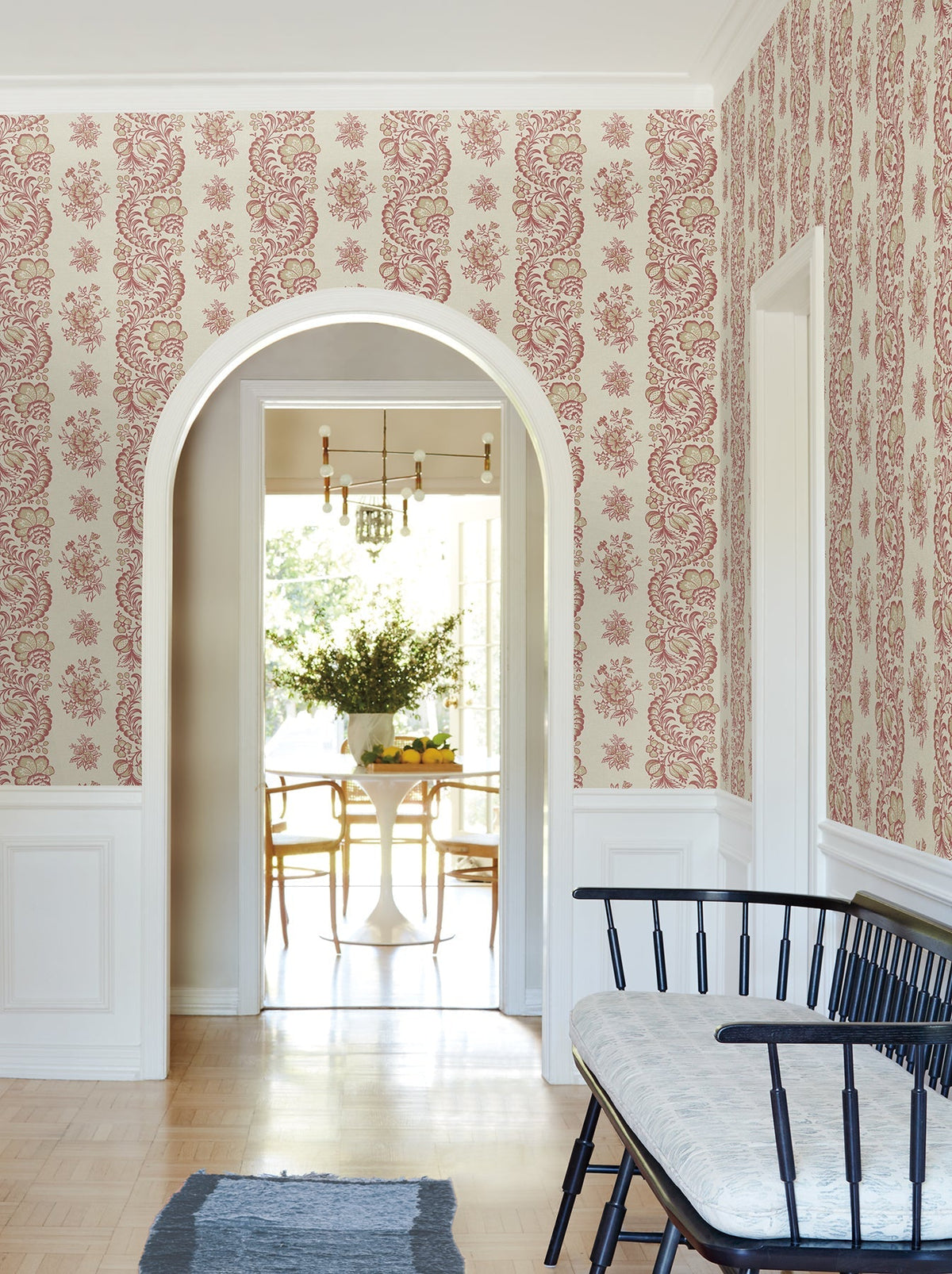 A hallway adorned with York Wallcoverings’ Beaumont Floral Stripe Wallpaper, featuring an arched doorway that opens to a dining area with a white table and chairs, and a black bench with a cushion set beside a white wainscoted wall, all illuminated by natural light.