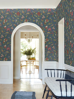 Room featuring Parterre Jacobean Wallpaper by York Wallcoverings, a white archway, and a light wood floor. A black bench with a gray cushion sits in the foreground, while through the archway is a white dining table with wooden chairs and a vase of greenery.