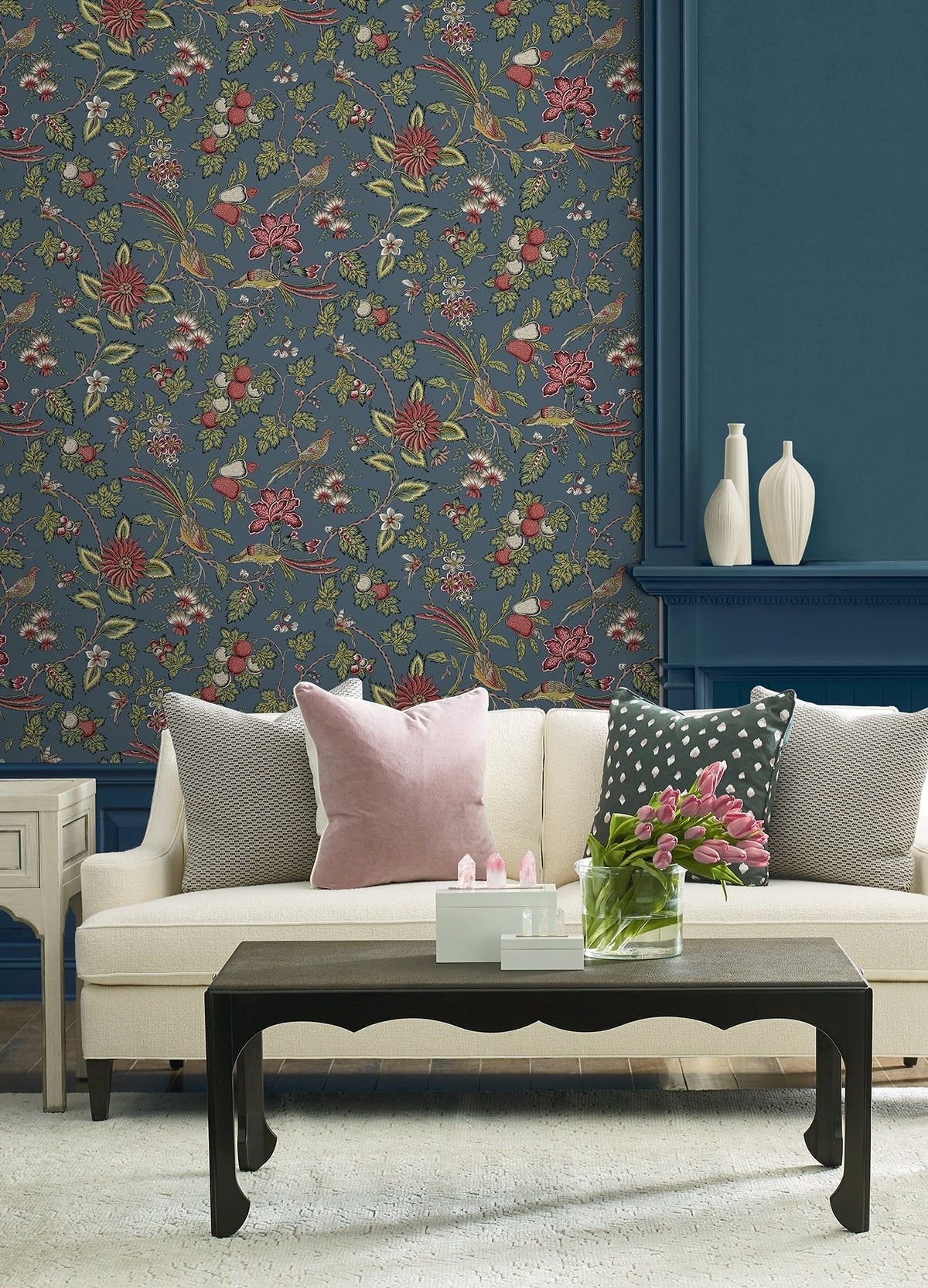 A cream sofa with assorted pillows sits in front of a blue Parterre Jacobean Wallpaper by York Wallcoverings. A black coffee table with pink tulips and white decorative vases is displayed, creating a cozy, stylish living room setting.