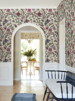 A room adorned with York Wallcoverings’ Parterre Jacobean Wallpaper, featuring white wainscoting and an arched doorway that opens to a dining area with a round table, chairs, a vase of flowers, and large windows filling the space with natural light. A black bench is positioned in the foreground.