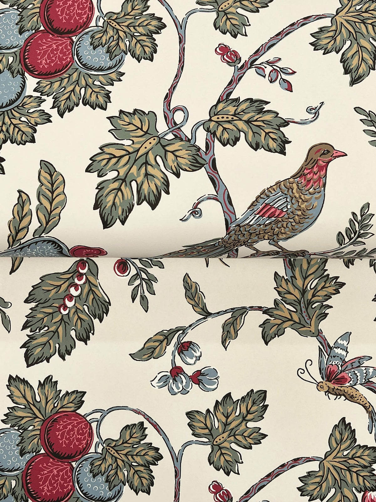 York Wallcoverings’ Parterre Jacobean Wallpaper showcases a nature-inspired pattern with birds, butterflies, leafy green branches, vibrant red and blue fruit, and delicate white berries set against a cream background.