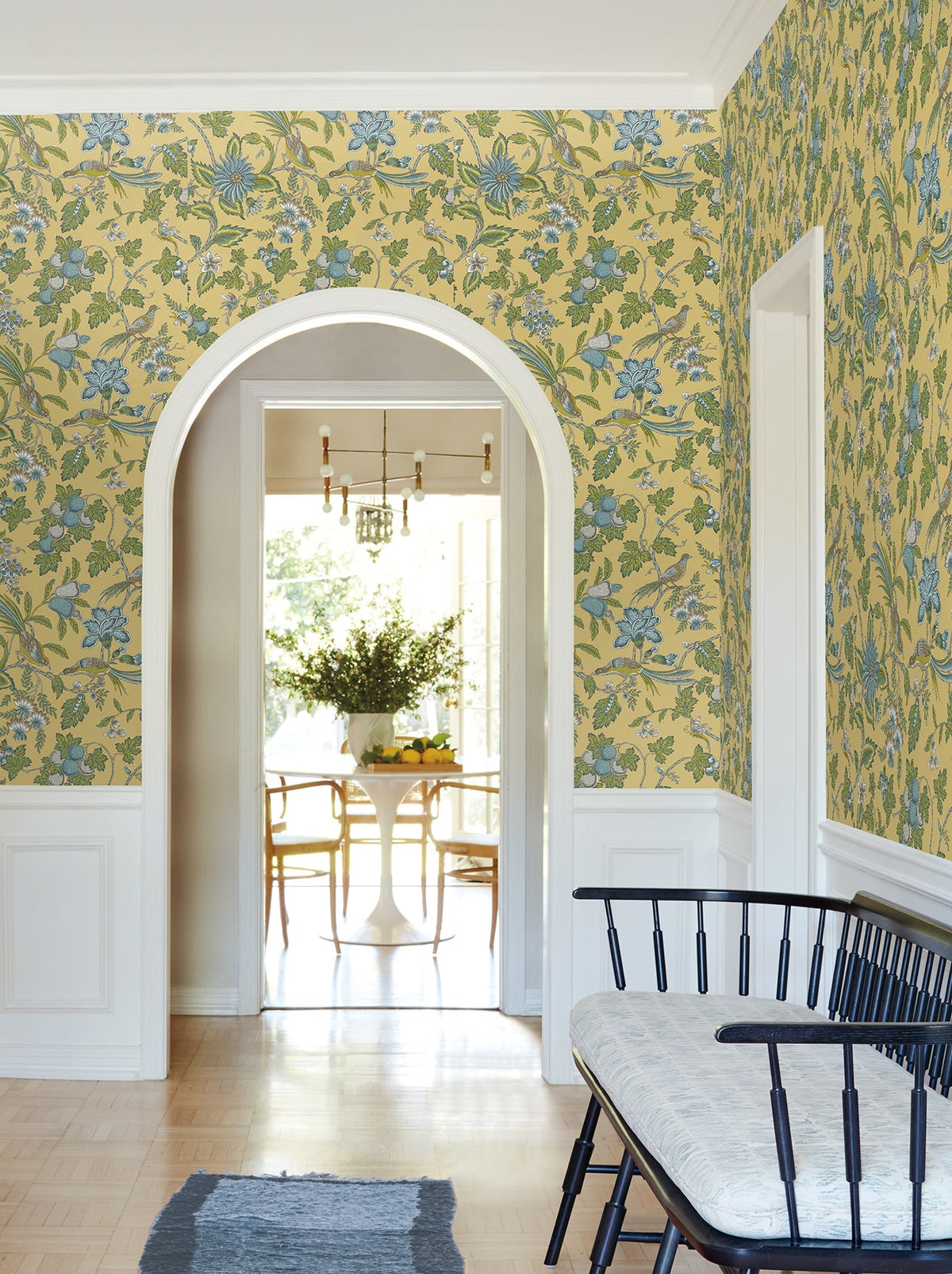 A hallway adorned with York Wallcoverings’ Parterre Jacobean Wallpaper in yellow and blue floral motifs, white wainscoting, a black bench topped with a cushion, and an arched doorway opening to a bright dining area furnished with a table and chairs.