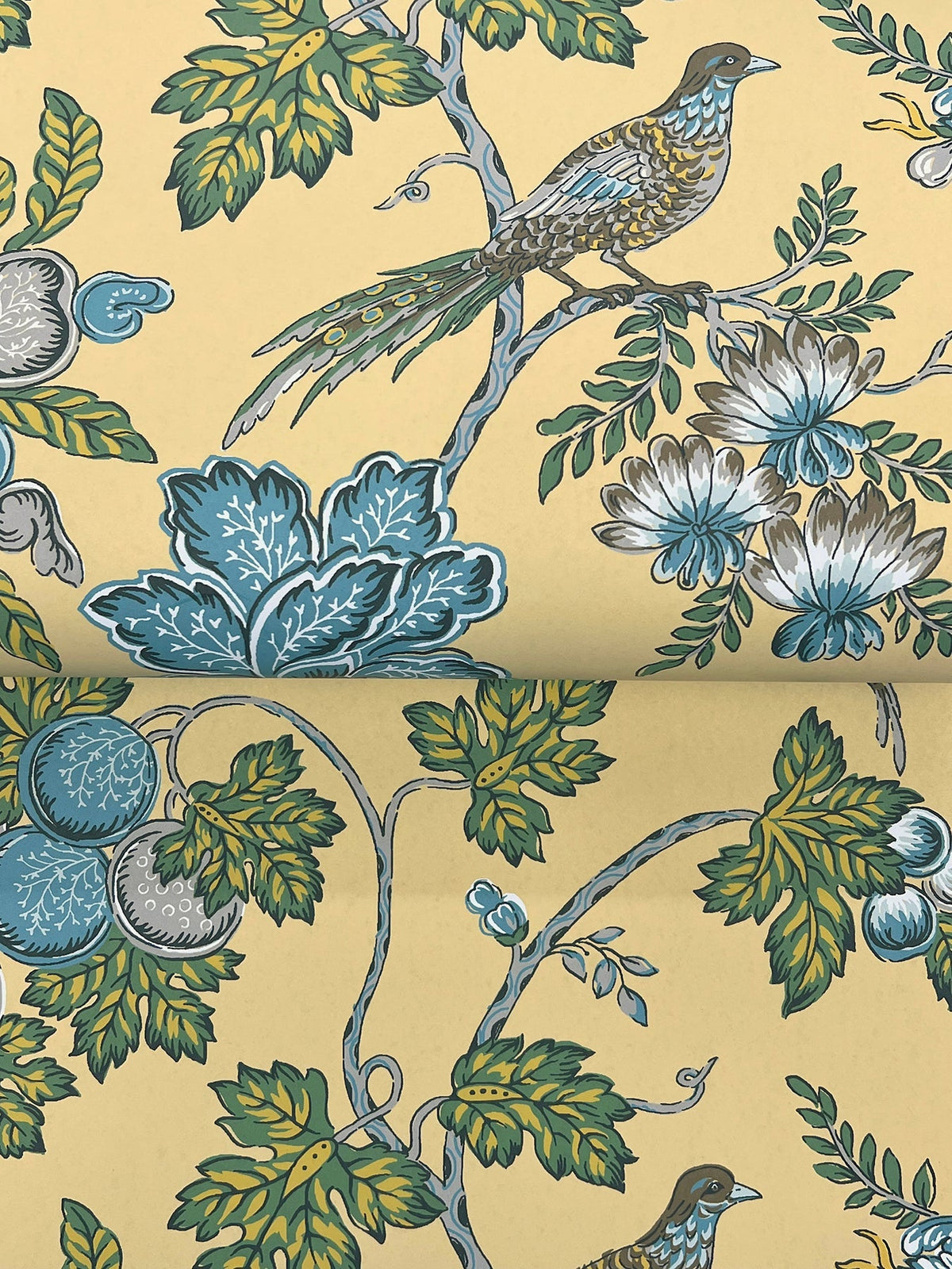 Introducing Parterre Jacobean Wallpaper by York Wallcoverings, featuring a repeating pattern of birds perched on branches amid blue-green leaves, white and blue flowers, and round blue fruits, all set against a yellow background.