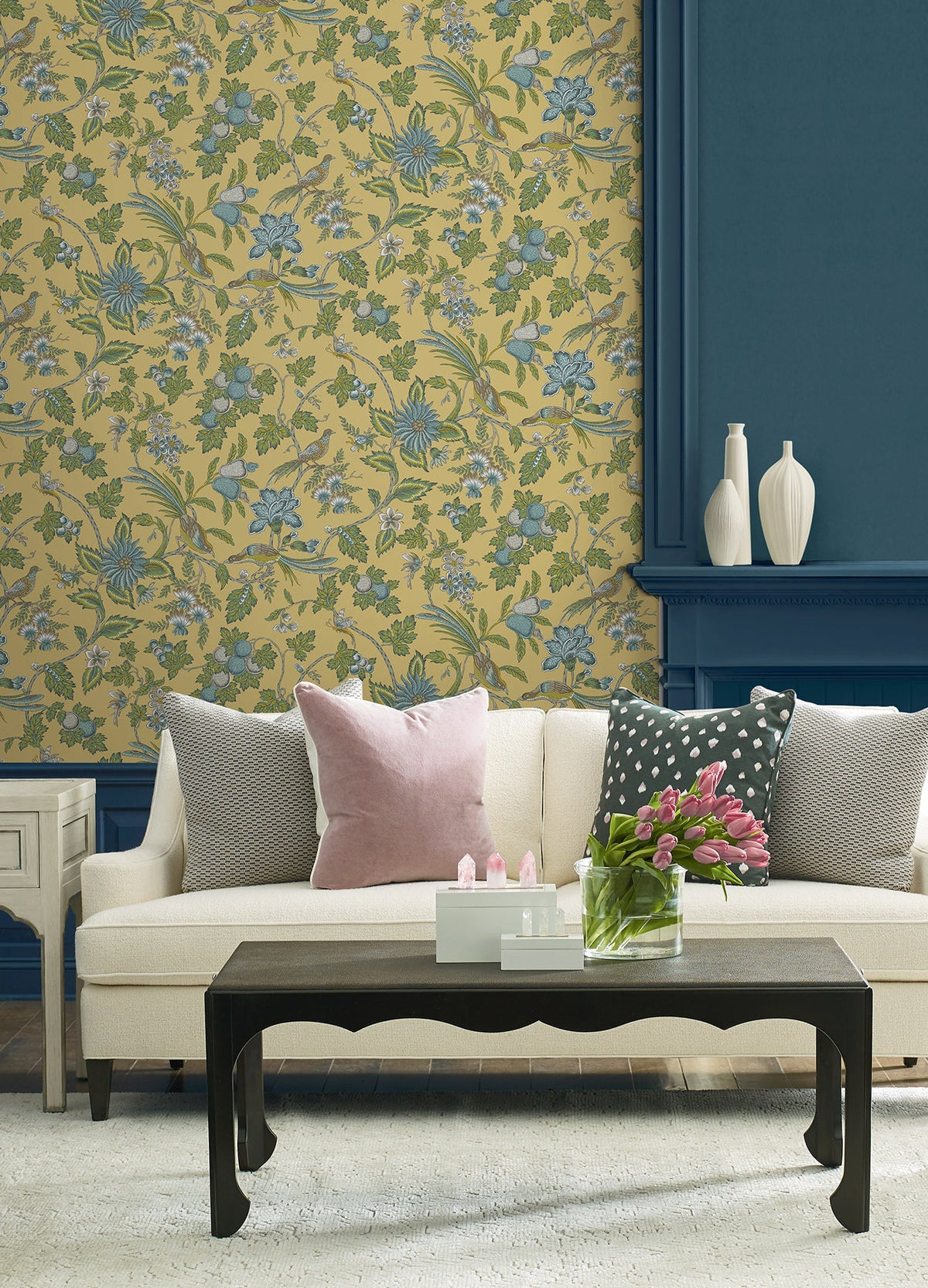 A cream sofa with throw pillows sits in front of York Wallcoverings’ Parterre Jacobean Wallpaper and a blue paneled wall. A black coffee table holds a glass vase with pink tulips, while three white vases are displayed on a shelf in the background.