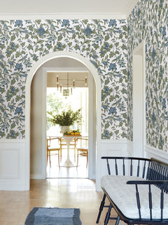 A hallway adorned with York Wallcoverings’ Parterre Jacobean Wallpaper in blue and green, featuring an arched doorway that opens to a sunlit dining room with a round table and chairs. A black bench with a cushion sits nearby atop a blue rug on the light wood floor.