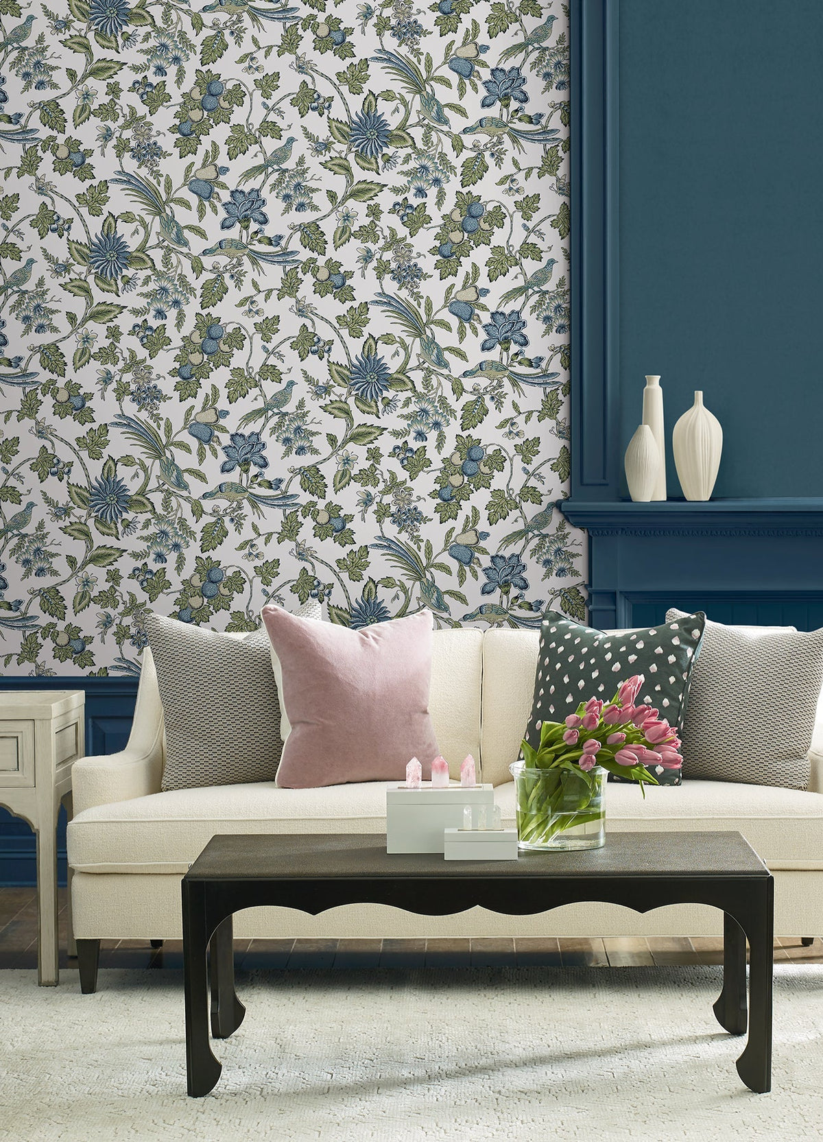 A cream sofa with pink and gray pillows sits in front of York Wallcoverings’ Parterre Jacobean Wallpaper. A black coffee table holds pink tulips in a glass vase, while white decorative vases are displayed on a blue shelf behind the sofa.